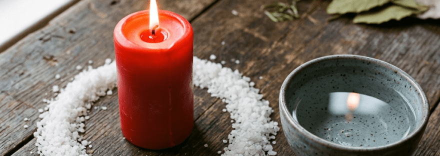 Red candle burning inside a circle of coarse white salt next to a ceramic bowl of water on a wooden surface