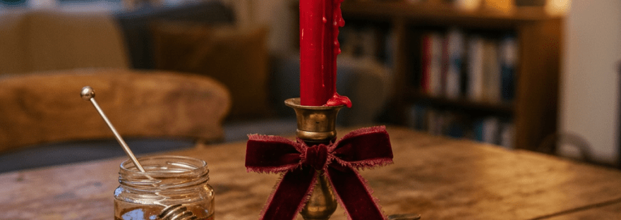 Lit red candle in brass holder with red bow, honey jar with dipper, and handwritten journal on wooden table